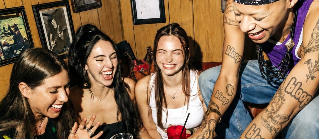 Quattro amiche che ridono attorno a un tavolino di un locale, accanto a un braccio tatuato Bloop