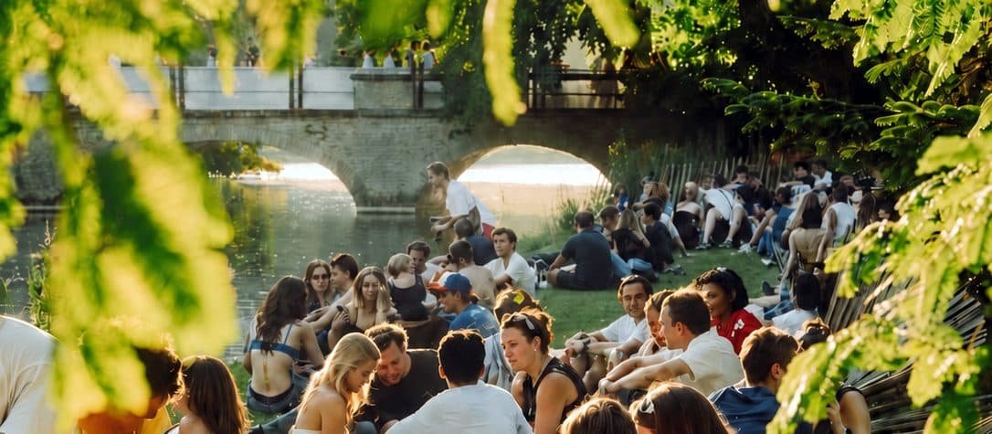 Un ponte sul canale con la riva affollata di gente seduta, inquadrato tra le foglie